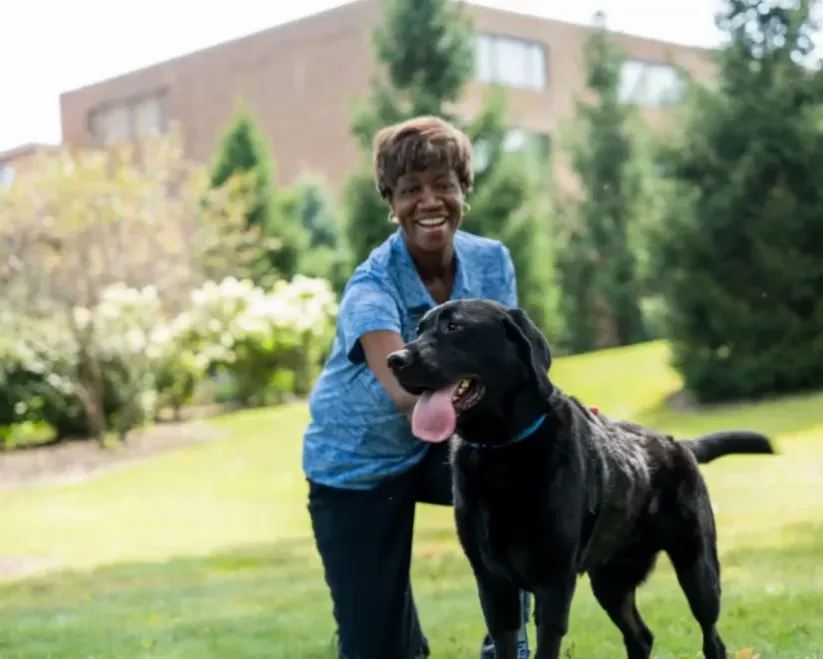 Mary Lester, owner of Pet Butler Tallahassee, FL is smiling with a dog who has his tongue out on the grass outside