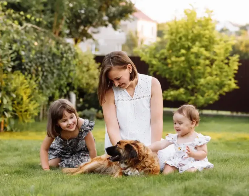 Woman sitting in the grass with a toddler girl and baby girl and petting a brown dog.