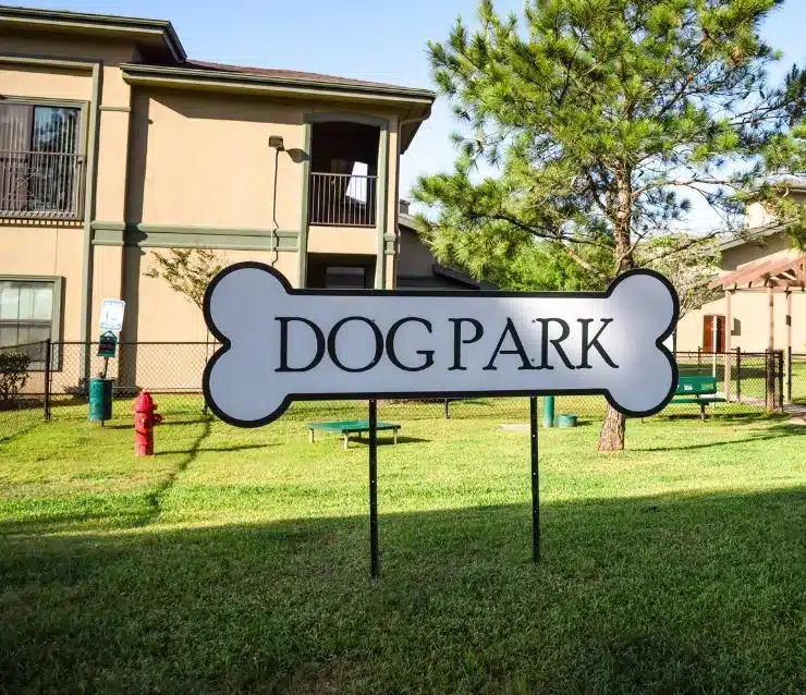 Large white dog park sign in the shape of a dog bone in green space outside of two-story multi-family buildings.