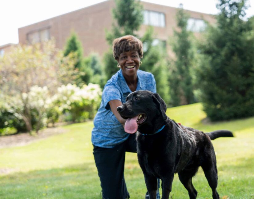 Mary Lester, owner of Pet Butler Tallahassee, FL is smiling with a dog who has his tongue out on the grass outside