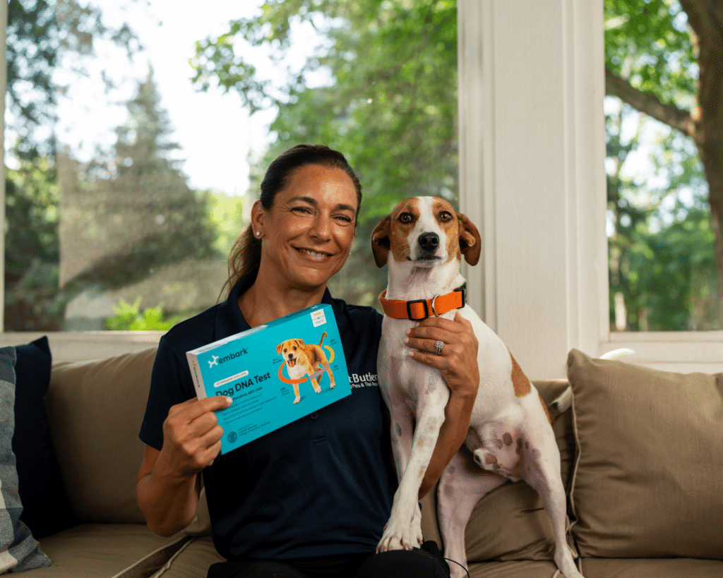 A smiling woman in a Pet Butler shirt holds a dog DNA test kit while sitting next to a white and brown dog wearing an orange collar on a cozy porch.