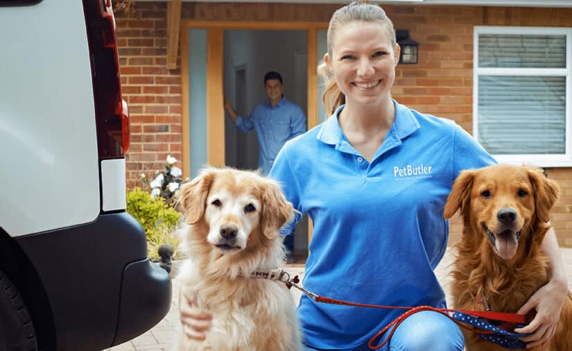 A smiling Pet Butler employee kneels with two happy dogs on leashes outside a home, while a man watches from the doorway.